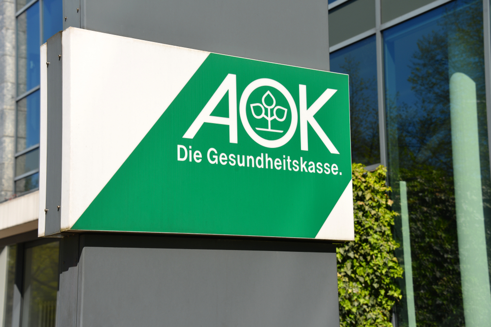 Ein Schild an einer Wand mit dem Logo der AOK Gesundheitskasse. Das Schild befindet sich links neben einem Gebäude der AOK.